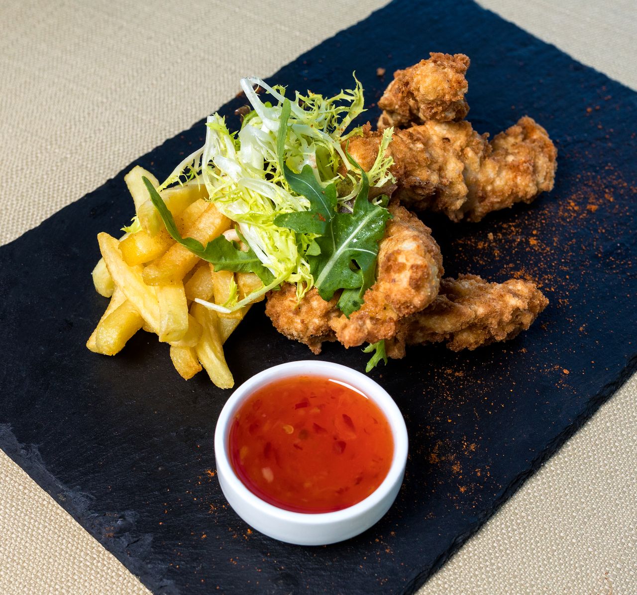Mouthwatering plate of fried chicken with crispy fries and fresh salad on a black slate, served with dipping sauce.